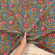 A close-up of a hand holding a piece of blue cotton fabric with a detailed floral block print in pink, red, and green, set against a rustic white wooden background.
