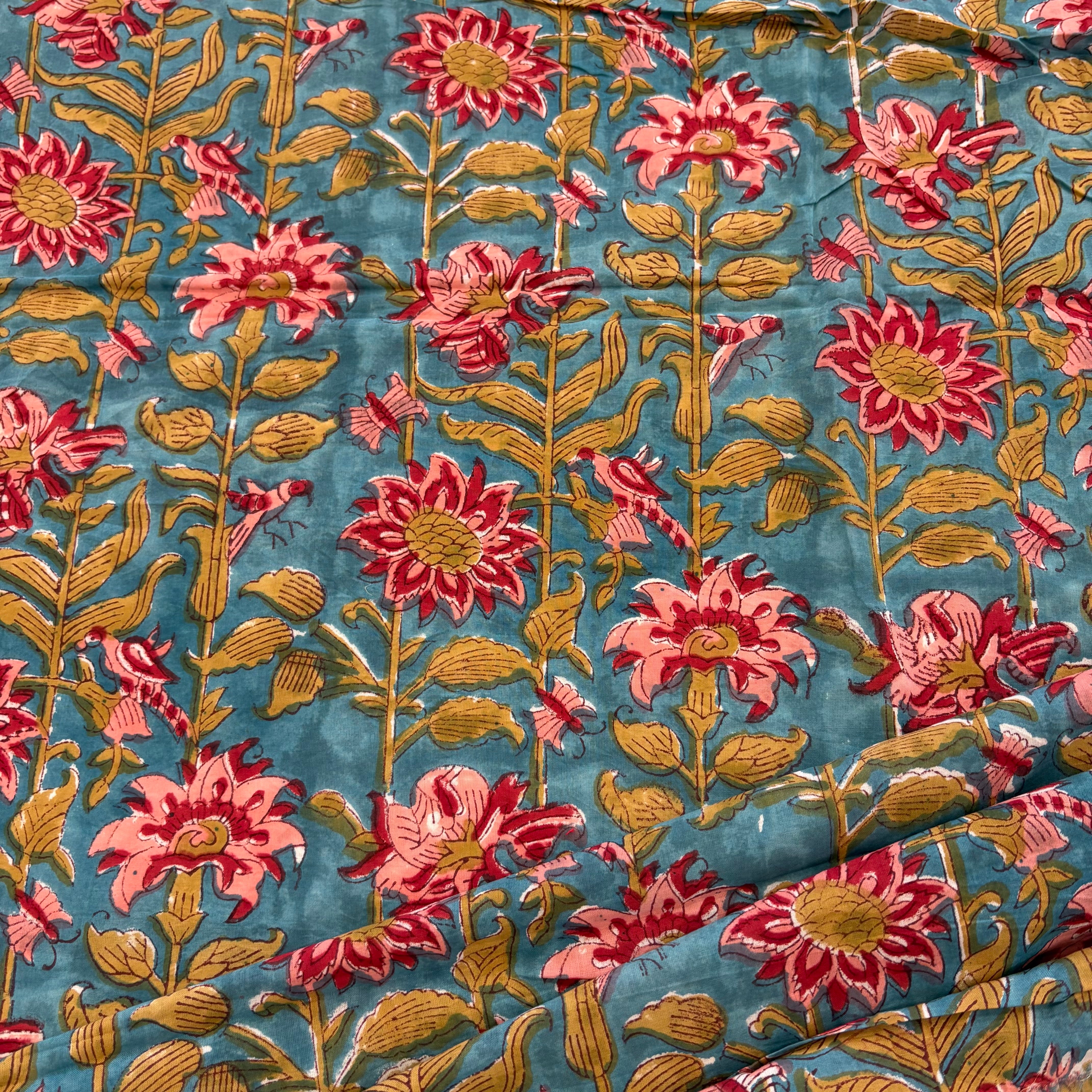 A close-up of a printed textile or fabric with a repeating floral pattern on a blue-green background. The flowers are pink to coral with green centers and stems, and the leaves are olive green.