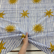 A hand pulling and gathering a piece of fabric with a pattern of vertical navy blue stripes and mustard yellow sun motifs on a white background, laid over a rustic white wooden surface.