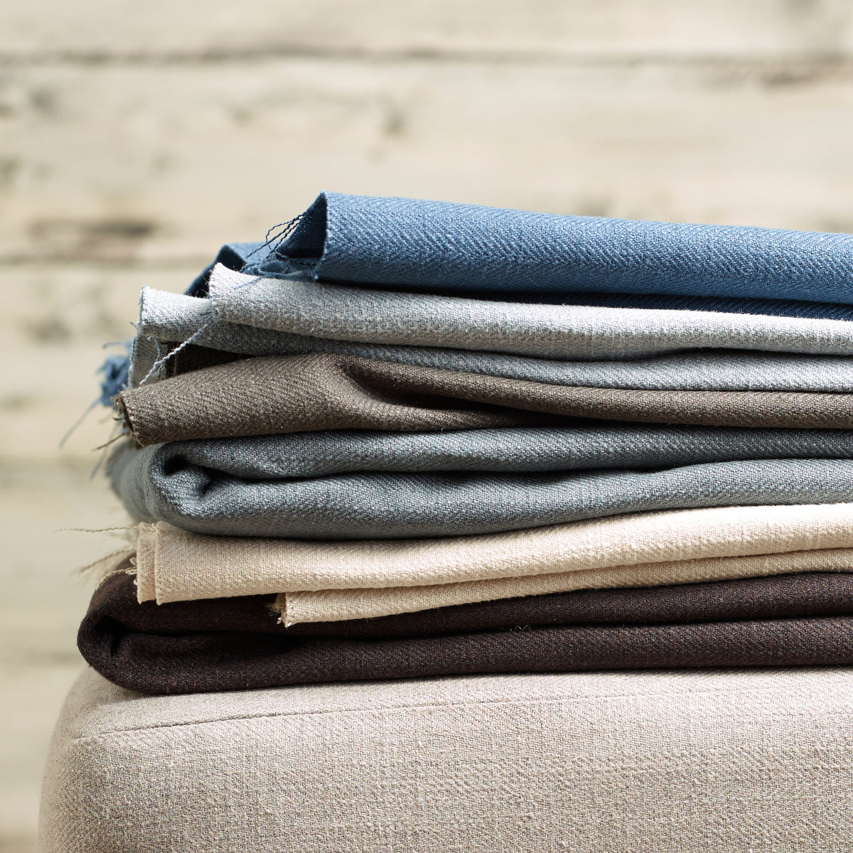 A stack of folded woven fabric swatches in blue, gray, taupe, cream, and dark brown, resting on a light beige upholstered surface against a softly blurred neutral background.