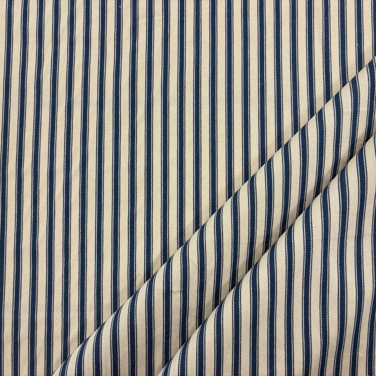 Classic navy blue cotton ticking fabric with traditional stripes and a soft handle folded at corner, ideal for cushions, tablecloths, table runners, lampshades, home furnishings and DIY craft projects. Timeless striped cotton fabric for contemporary and country style interiors.