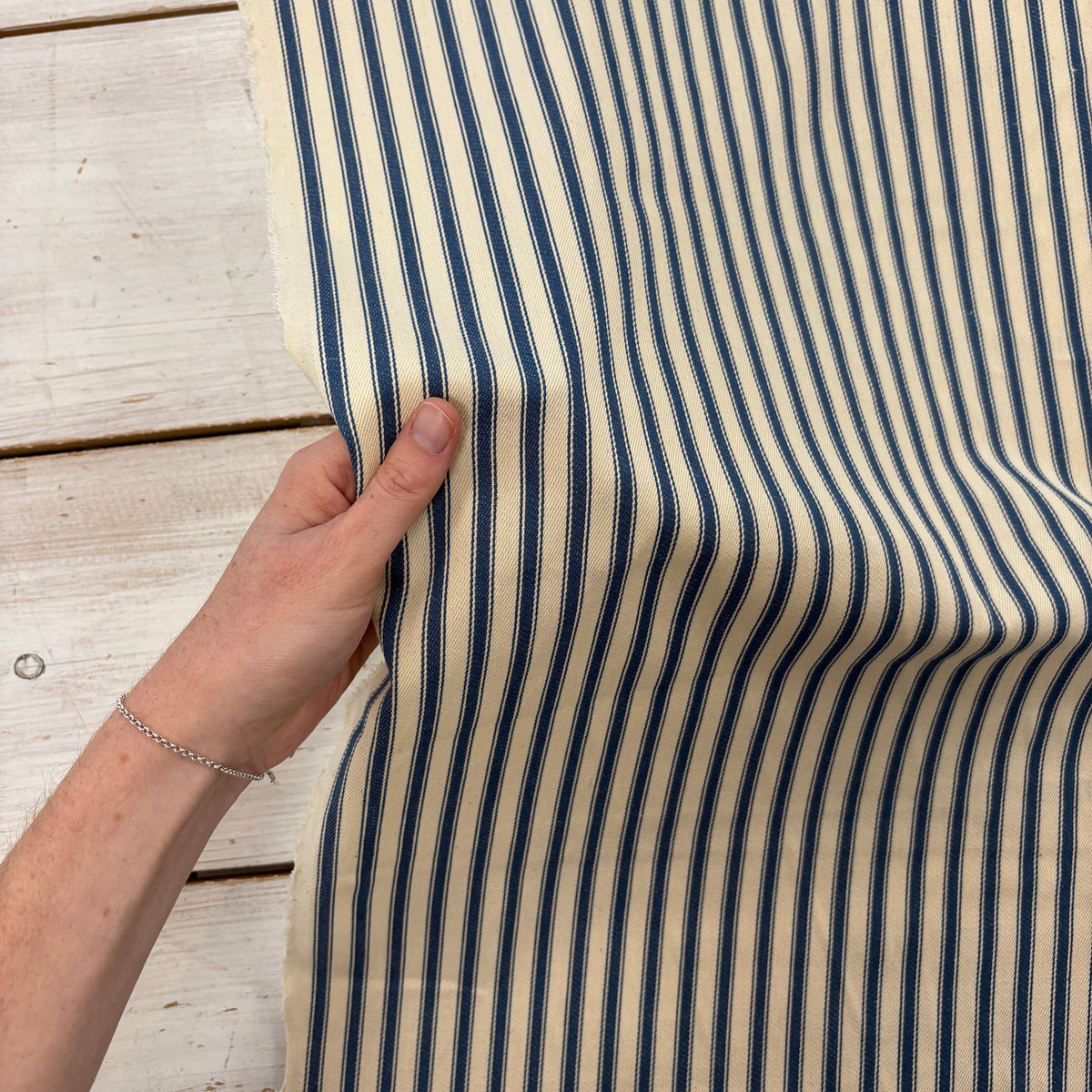 Classic navy blue cotton ticking fabric with traditional stripes and a soft handle with hand for scale, ideal for cushions, tablecloths, table runners, lampshades, home furnishings and DIY craft projects. Timeless striped cotton fabric for contemporary and country style interiors.