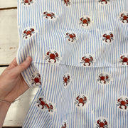 A close-up of a hand holding a piece of fabric with a pattern of blue and white vertical stripes and red crabs on a white background, laid over a white wooden surface.