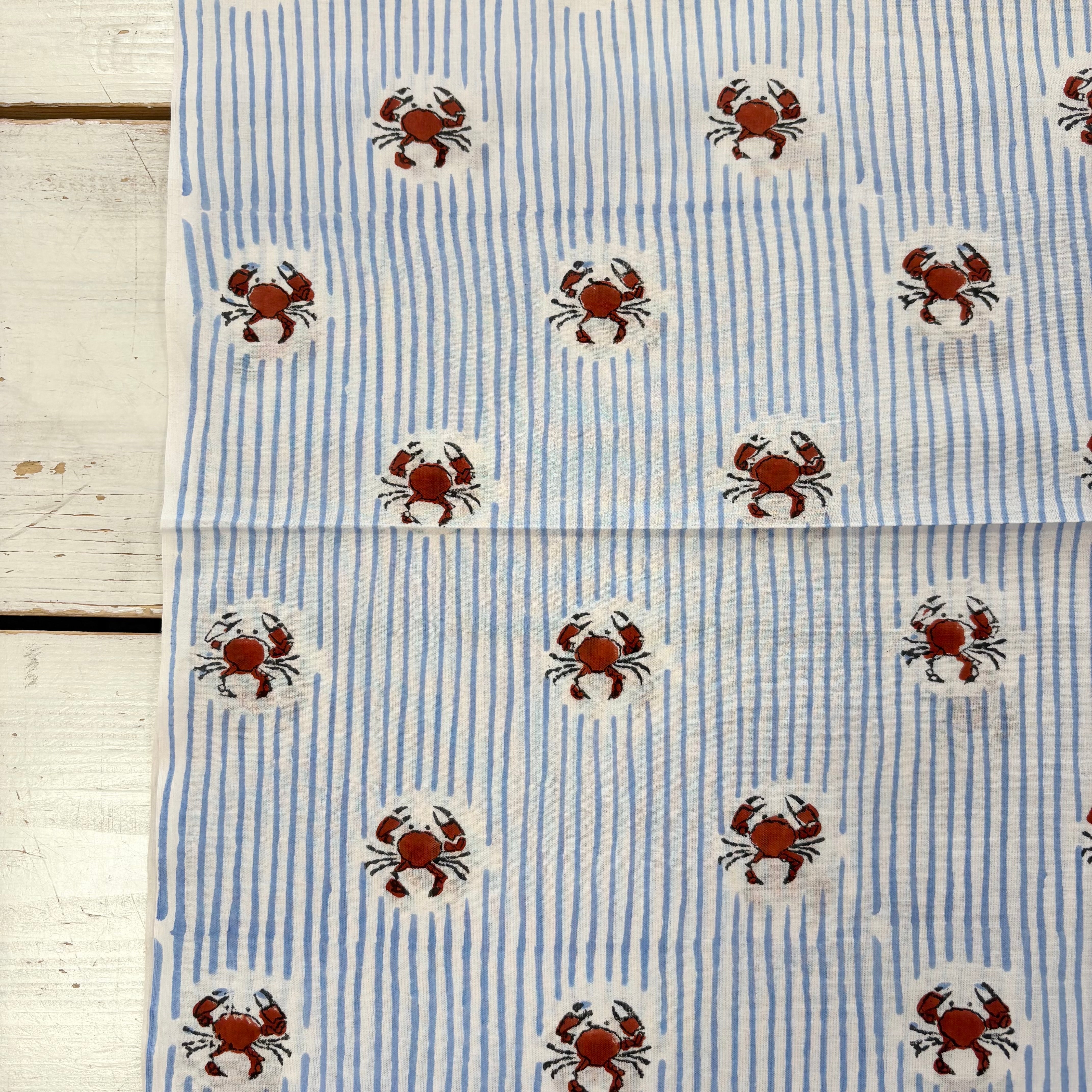 Blue and white striped fabric with a repeating pattern of brown crabs on white circular backgrounds, laid flat on a white wooden surface.