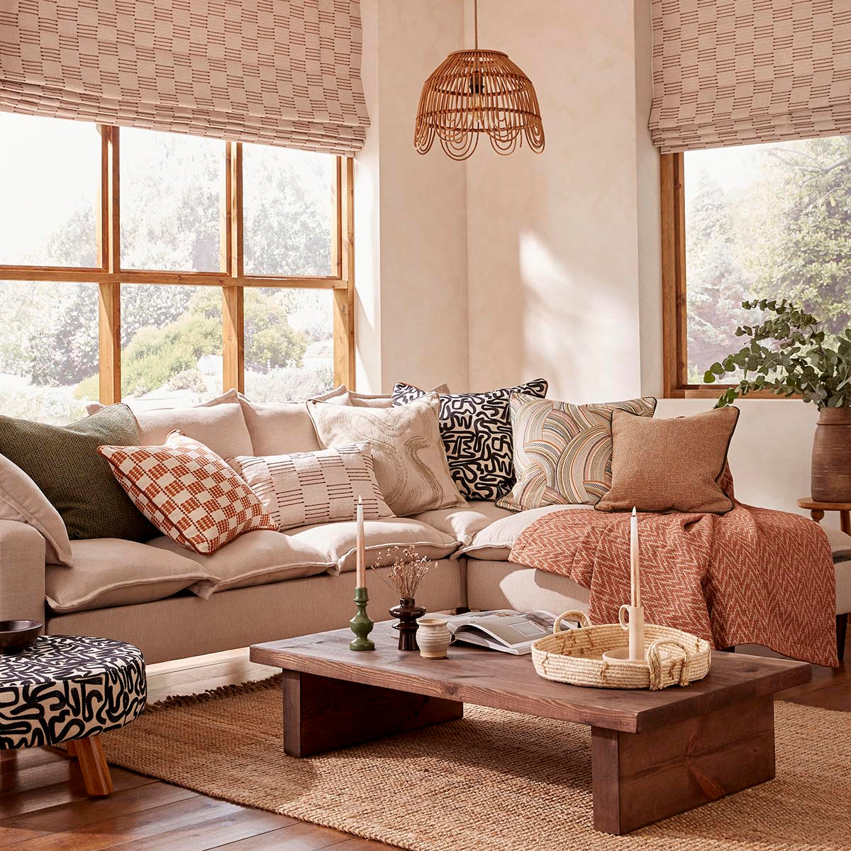 A styled living room featuring a beige sectional sofa with multiple patterned throw pillows, a woven wicker pendant light, and two windows with beige checkered Roman shades.