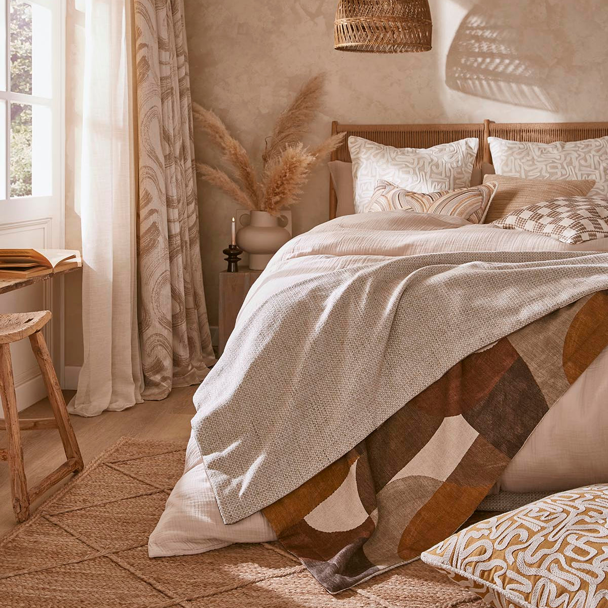A cozy bedroom scene with a bed dressed in neutral-toned bedding, including a cream duvet and patterned pillows, styled with a woven pendant light, textured wallpaper, and a window with sheer curtains.