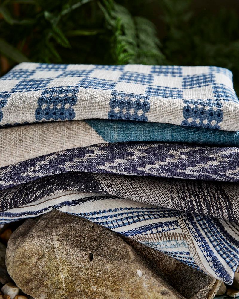 A stack of folded woven textiles, likely towels or blankets, featuring blue and off-white patterns, displayed outdoors on a stone with blurred green foliage in the background.
