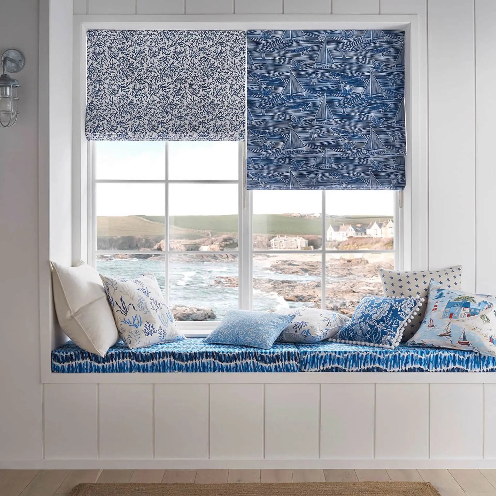 Nautical-themed window seat set with blue patterned cushions and matching roller blinds in a bright, white-paneled room overlooking the sea.