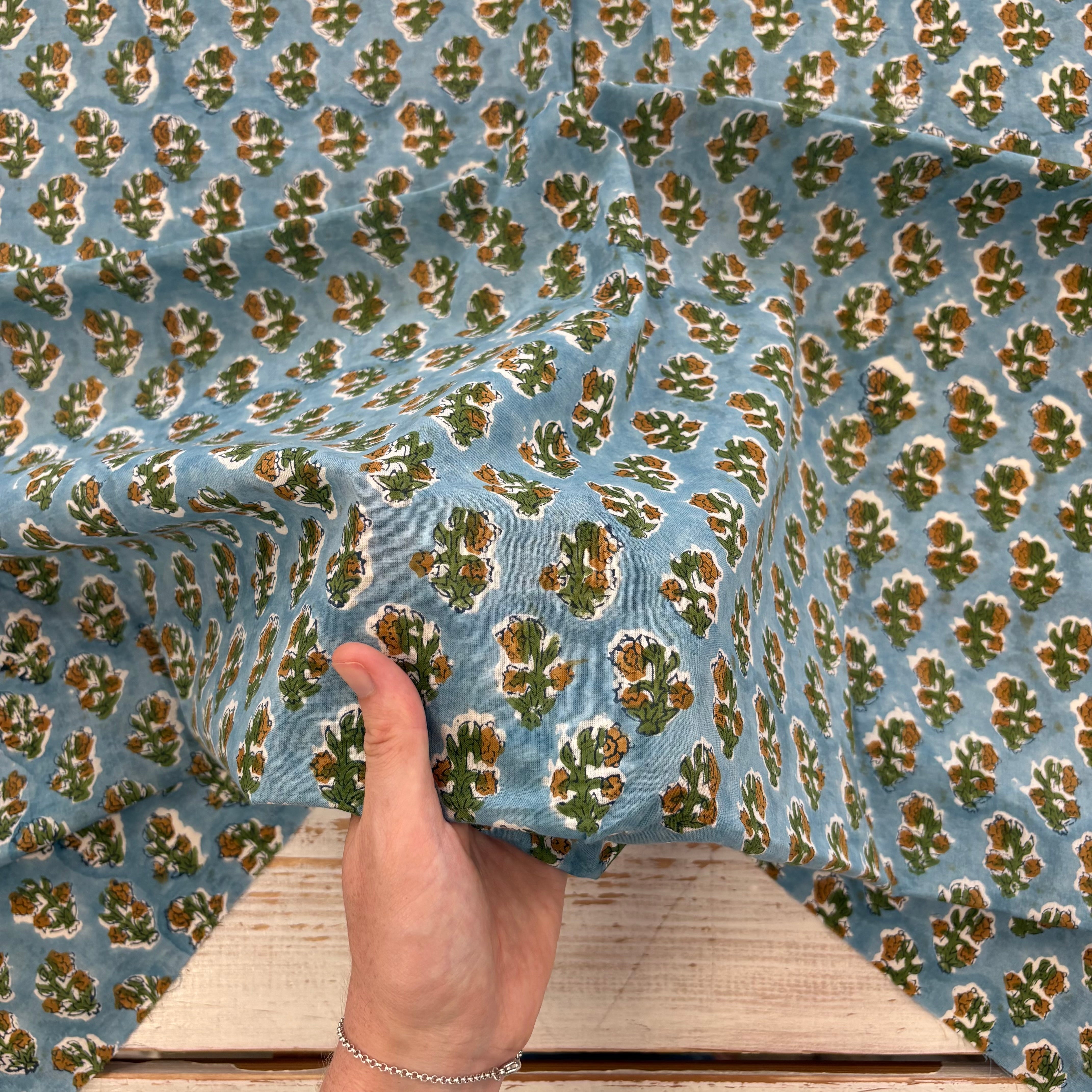 A close-up of a light blue fabric with an all-over repeating green and mustard floral block-print pattern, held by a hand to show its drape and texture.