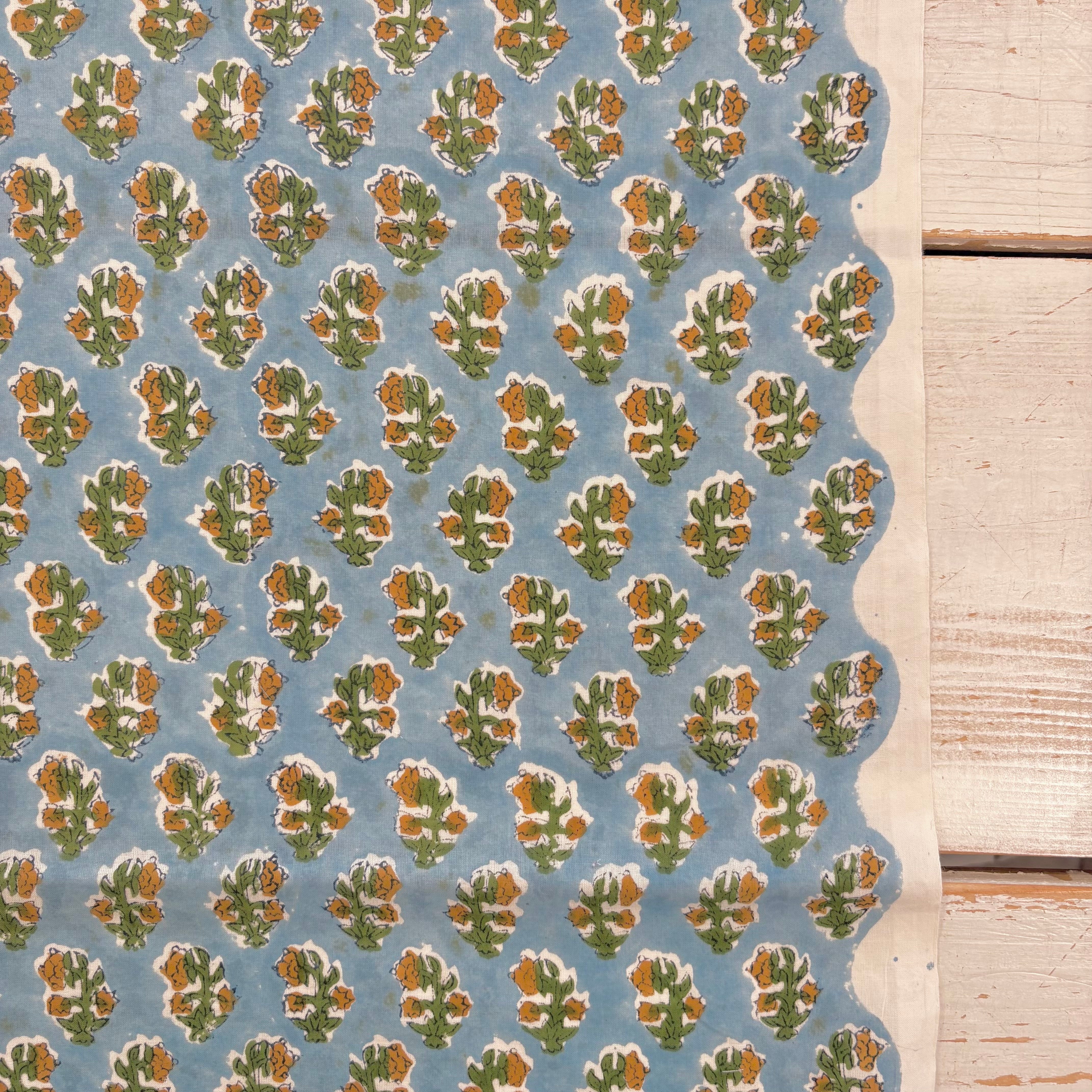 A close-up of a patterned fabric sample with a scalloped edge, featuring a repeating floral block print in green, orange-brown, and white on a dusty blue background, laid next to a light wood plank surface.