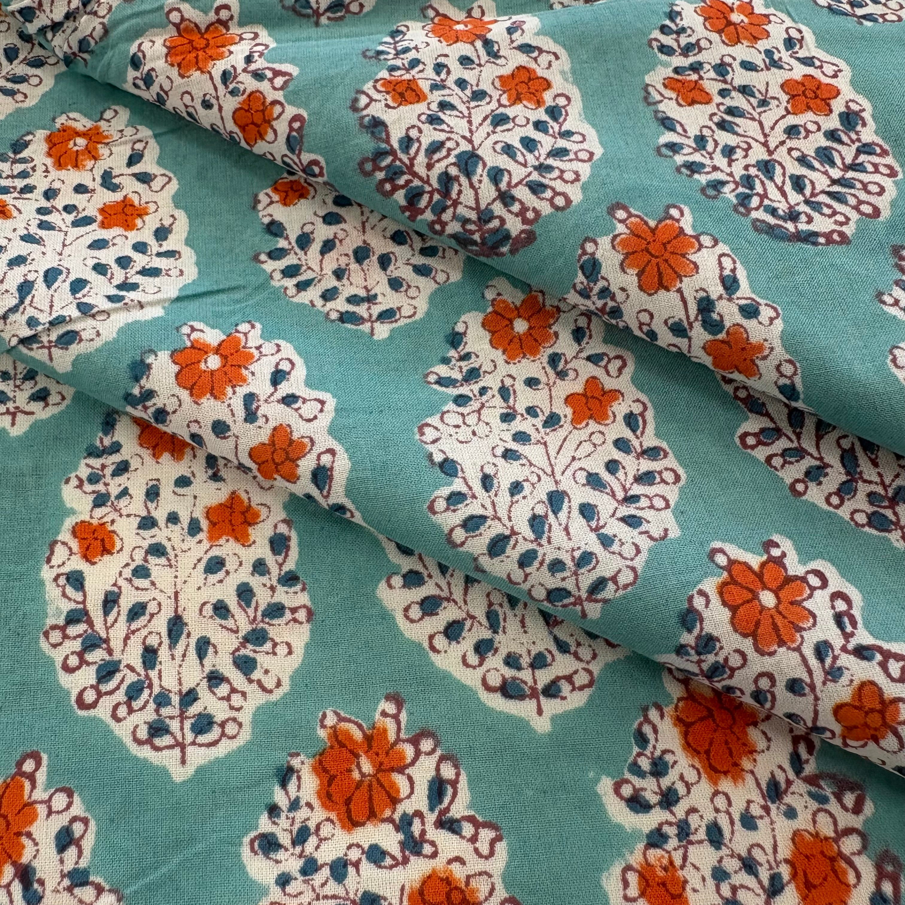 A close-up of patterned fabric with a teal background and repeating white floral motifs accented with orange and dark blue details.