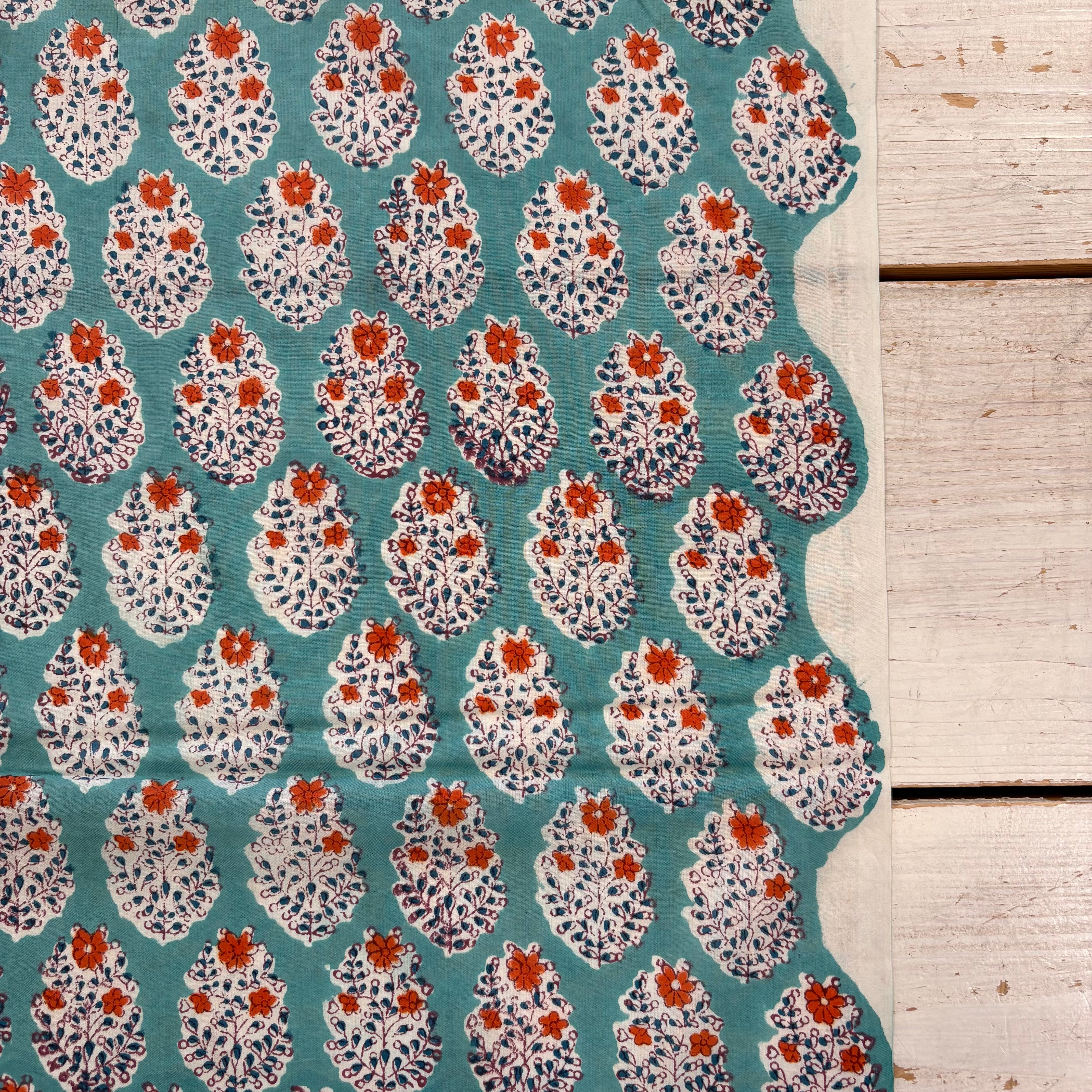 A close-up of patterned fabric with a teal background and repeating white floral or leaf-like motifs accented with orange and dark blue details, laid beside a light wood surface.