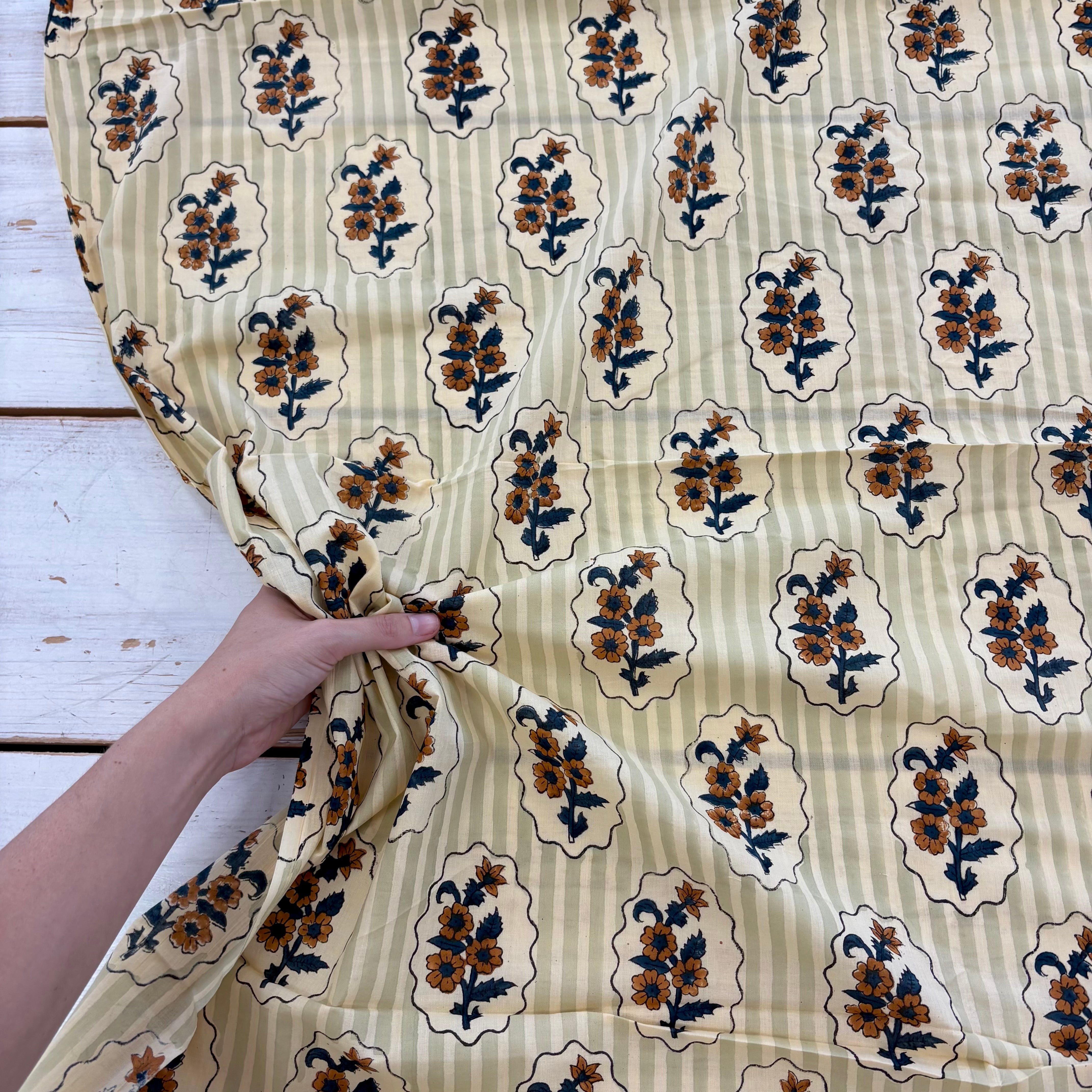 A hand holding a cream-colored cotton fabric with a vertical stripe pattern and a repeating block print of floral motifs inside decorative cartouches.