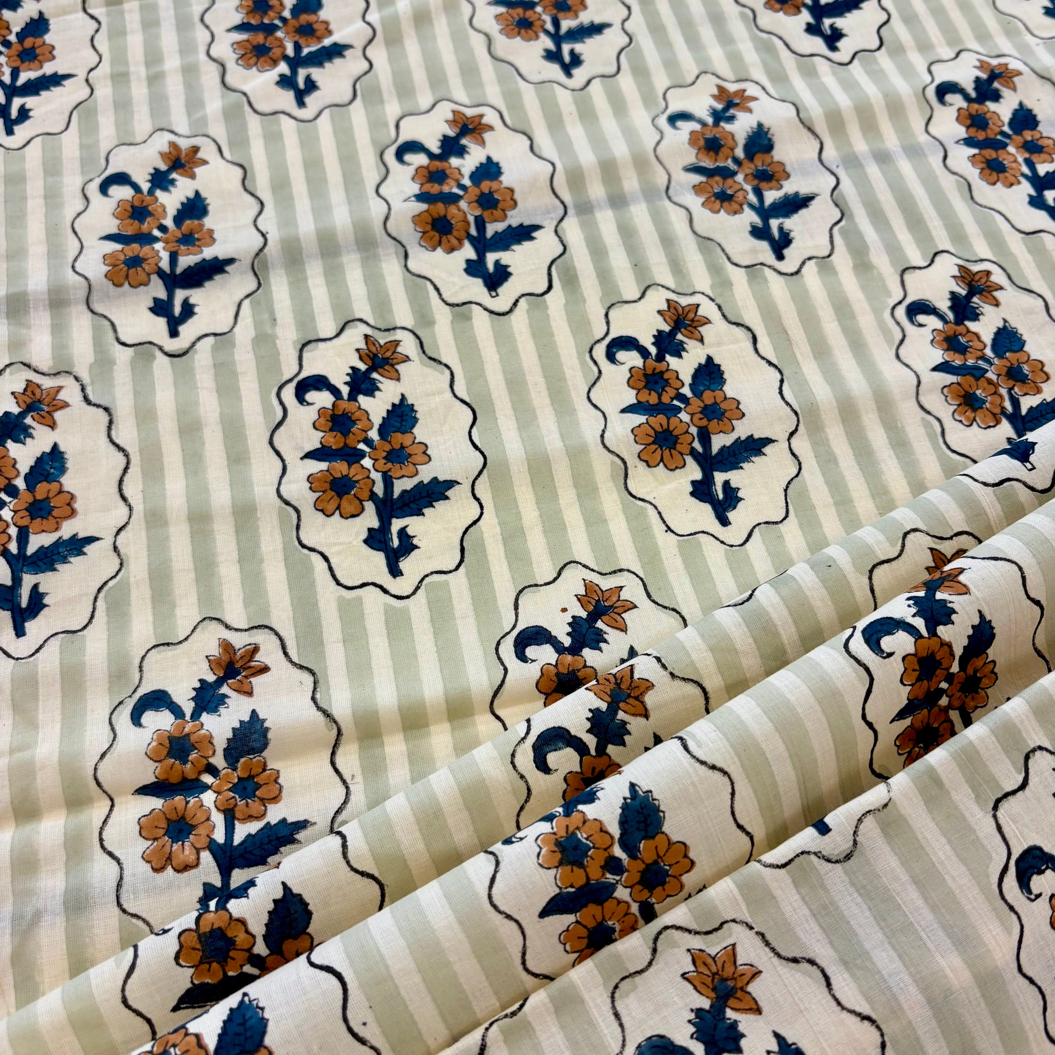 A close-up of patterned fabric with a cream and light sage green striped background, decorated with repeating floral medallions in blue, mustard yellow, and brown.