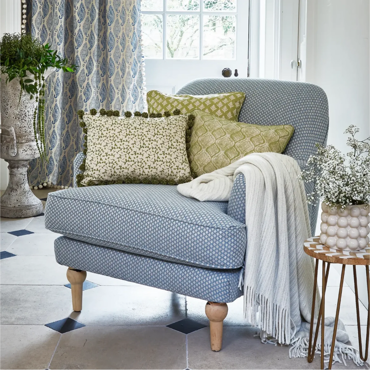 A blue and white patterned upholstered armchair in a bright living room setting, styled with green patterned and floral throw pillows, a white fringed throw blanket, and a small side table nearby.