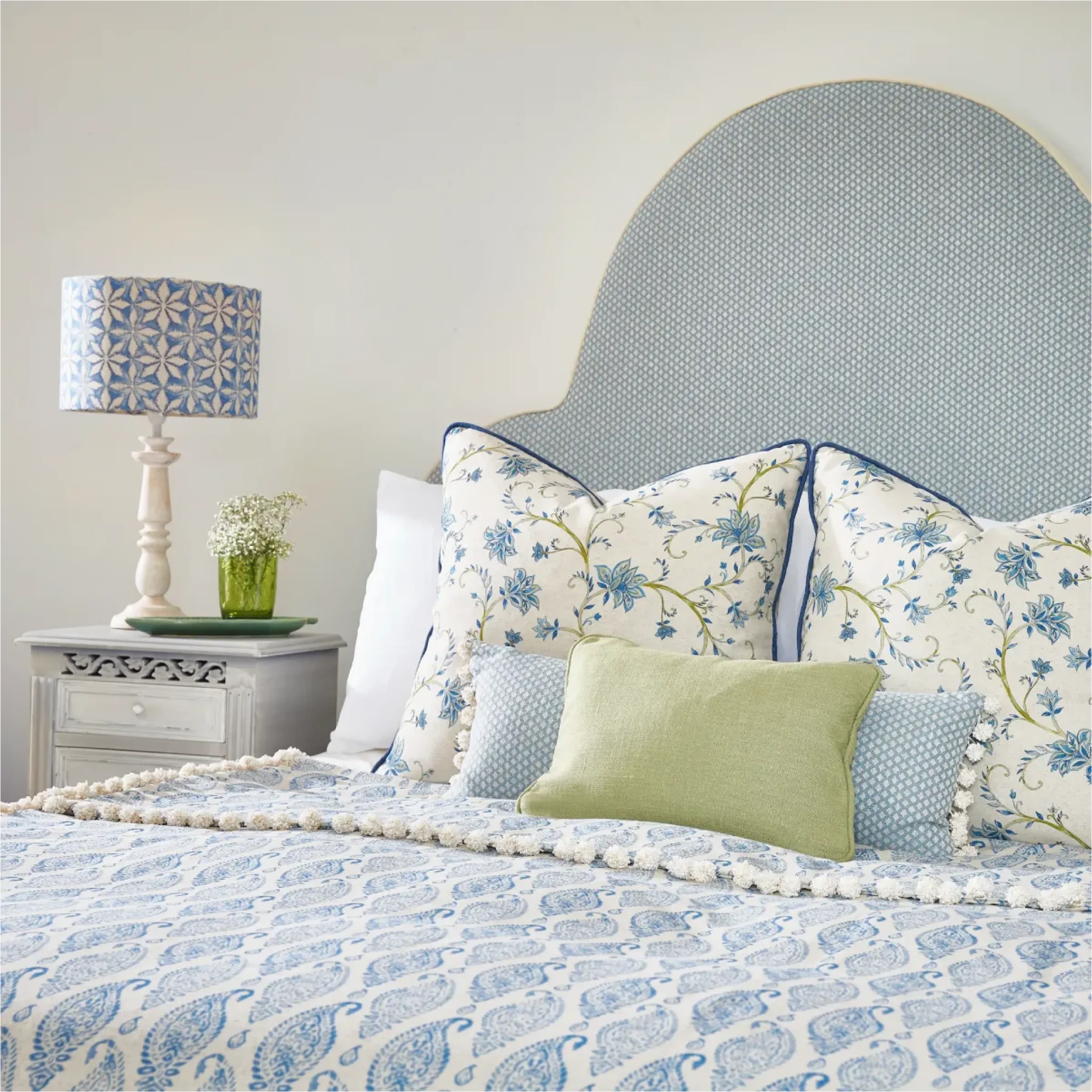 A styled bedroom scene featuring a bed dressed in a blue and white paisley patterned bedding set with matching patterned pillows, a light green accent pillow, and a large arched upholstered headboard. A white nightstand with a blue-and-white patterned lamp and a small vase is visible on the left.