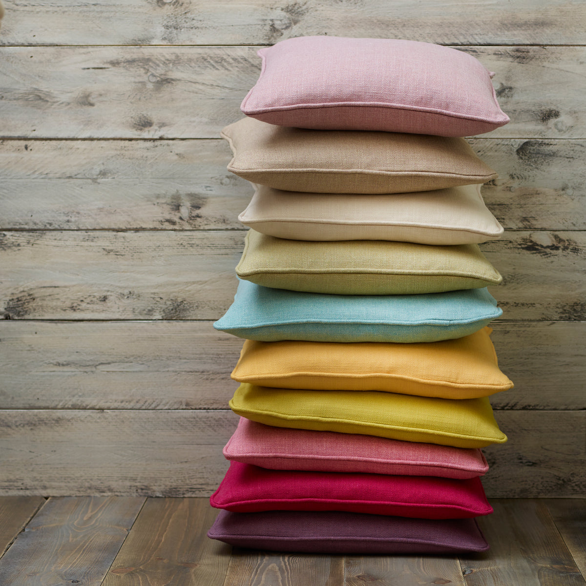 A stack of ten square throw pillows in various pastel and bright colors, including pink, beige, yellow, teal, orange, magenta, and purple, arranged on a rustic wooden plank floor against a matching wooden wall.