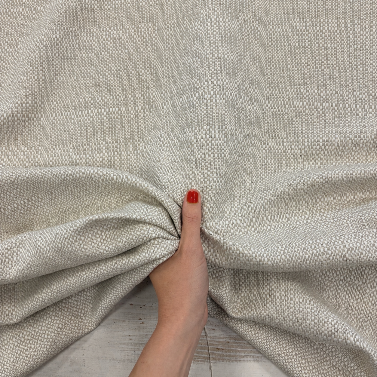 A close-up of a textured fabric in a neutral linen tone, gathered by a hand with red nail polish, showing the material's weave and drape.