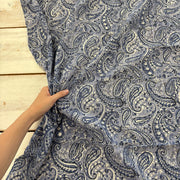 A hand holding up a piece of fabric with a blue paisley pattern, laid over a light wood plank surface.