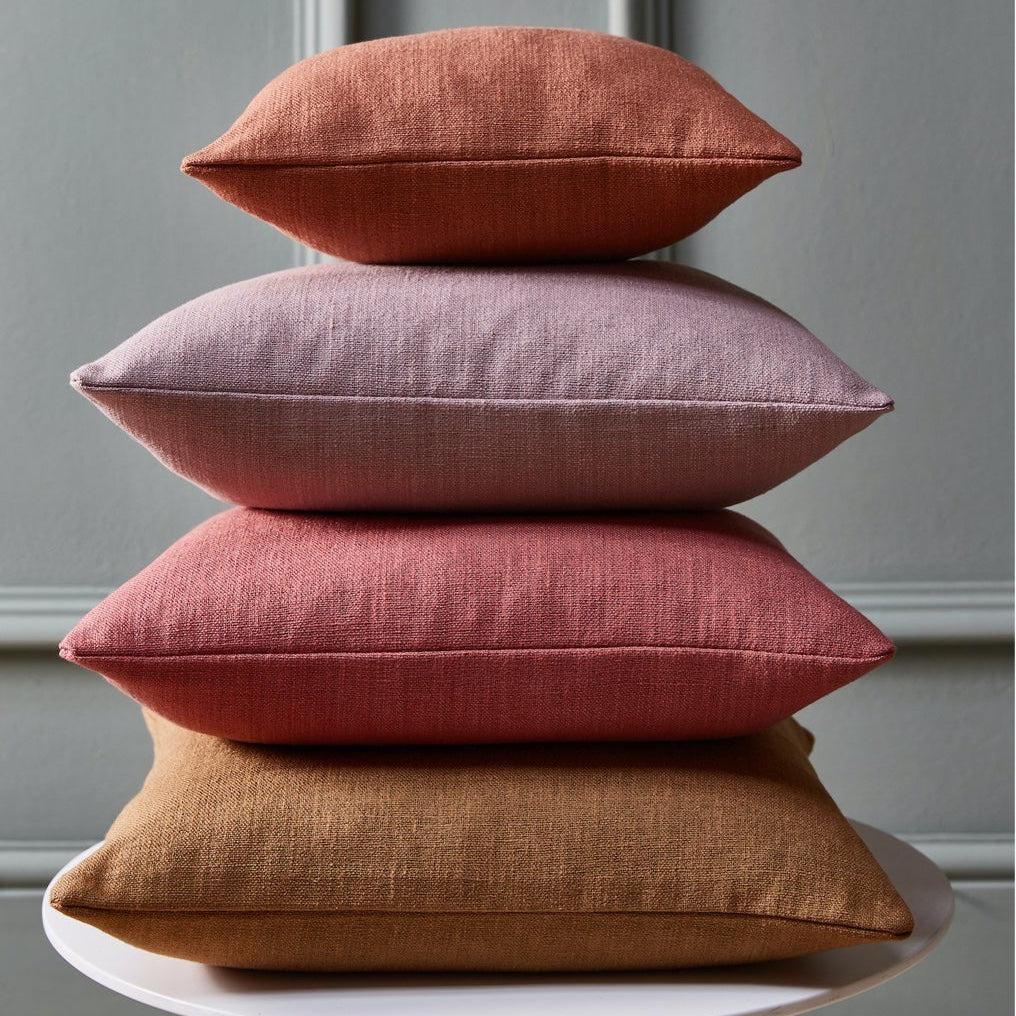 Four textured throw pillows in terracotta, dusty pink, muted red, and ochre brown stacked vertically on a white round table against a gray paneled wall.