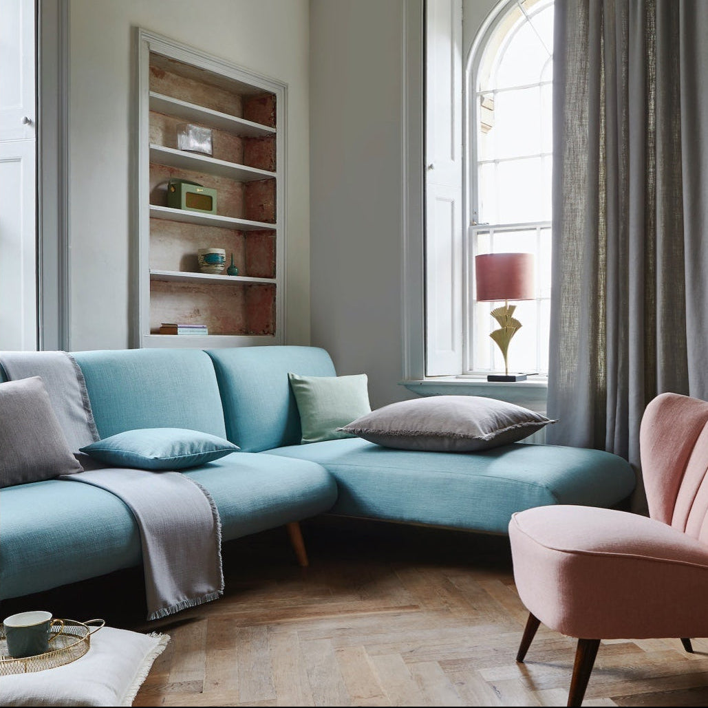 A modern living room featuring a light blue modular sectional sofa with gray and mint pillows, a dusty pink armchair, a built-in wooden shelf, and a large arched window with gray curtains.