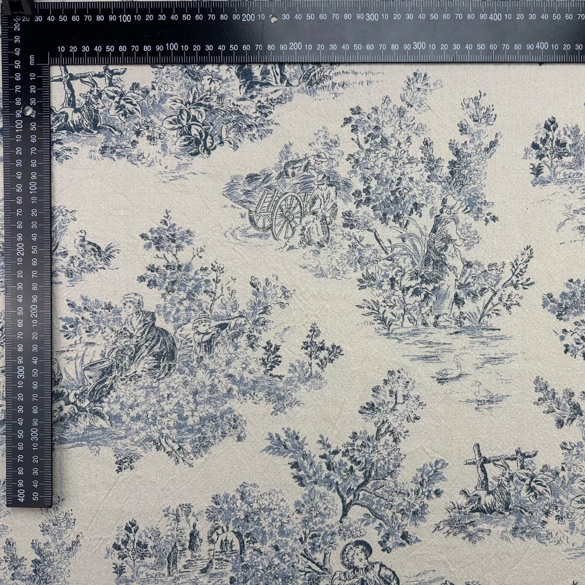 Rustic Toile Navy H412 Blue floral and landscape pattern on beige fabric with a ruler for scale.