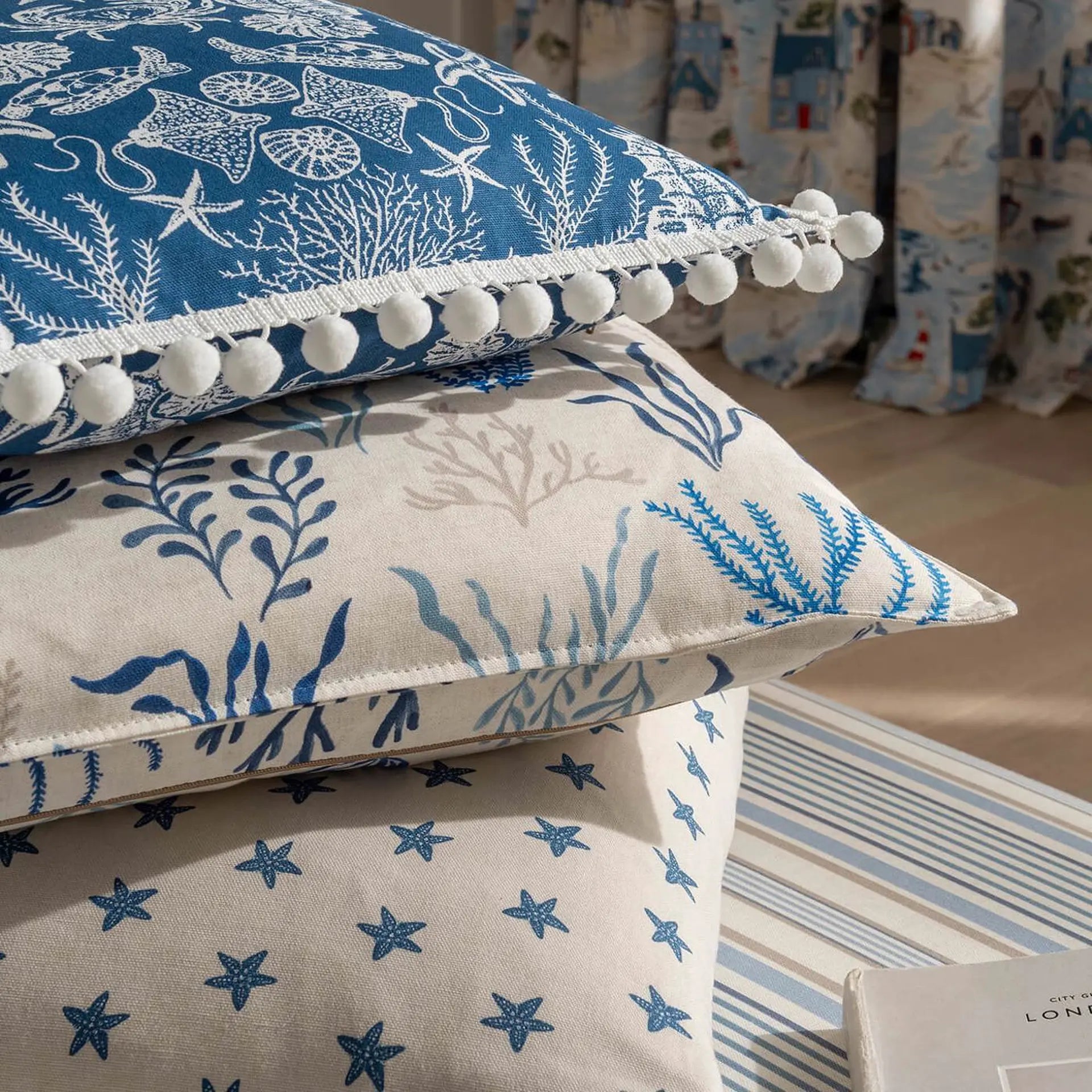 Three decorative throw pillows stacked on a striped surface, featuring nautical designs including starfish, sea urchins, and coral in blue and beige tones. The top pillow has white pom-pom trim along the edge.