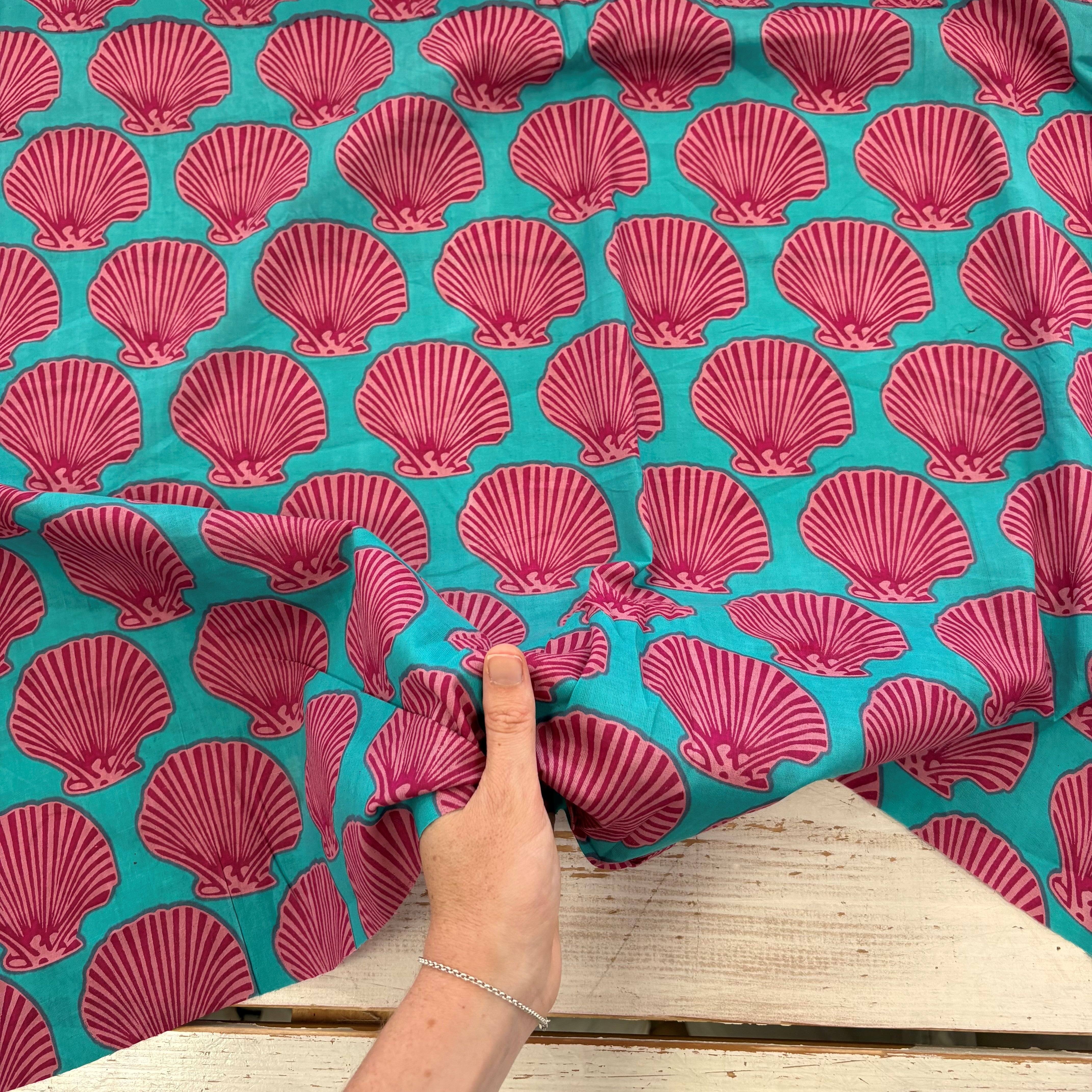 A close-up of a turquoise fabric with a repeating magenta-pink scallop shell pattern, held by a hand over a light wood surface.