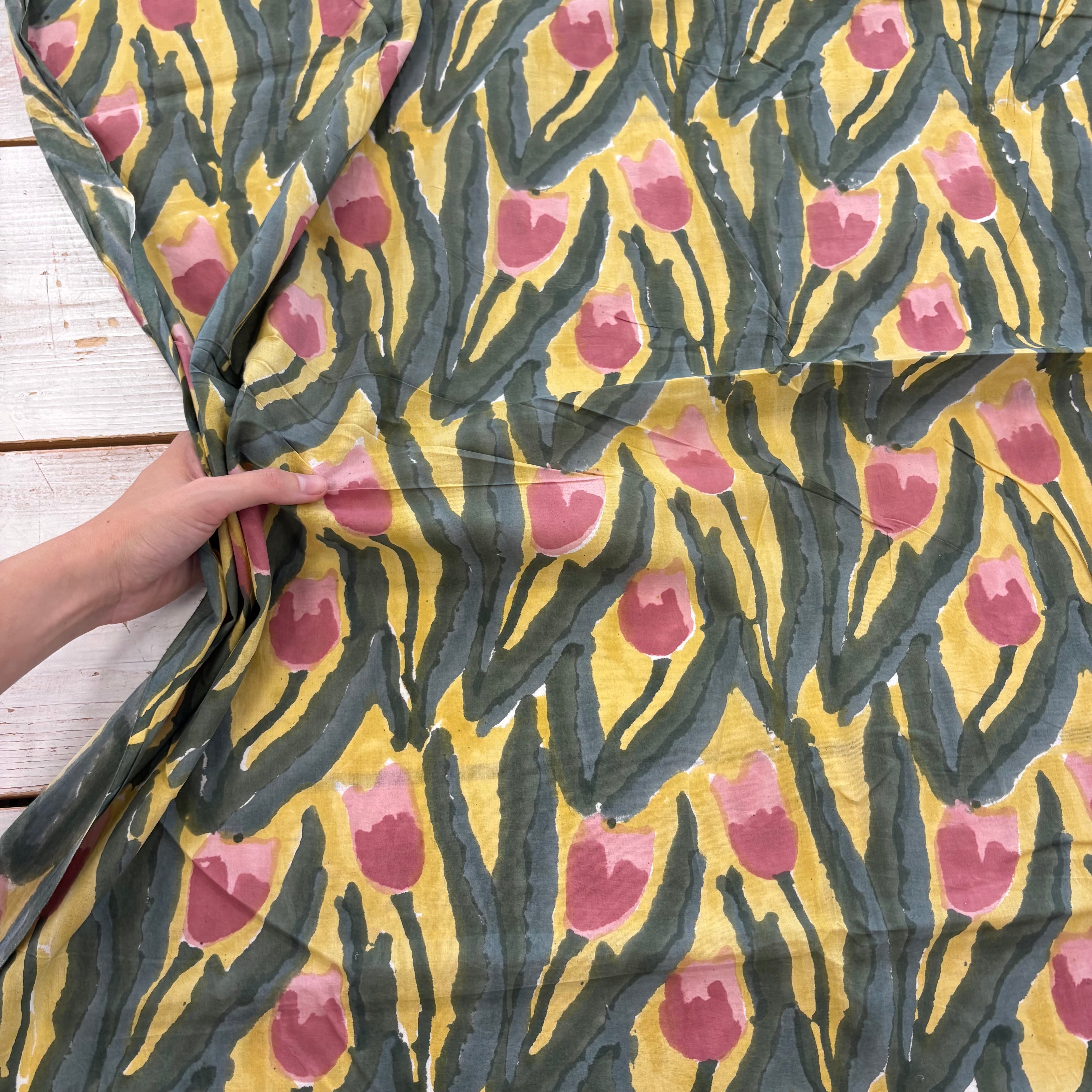 A close-up of a patterned fabric with a hand holding the side to show its drape. The fabric features a yellow background with dark green abstract leaf-like vertical shapes and pink tulip motifs.
