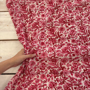 A close-up of a red and pink floral fabric with a hand holding the corner to show its drape and texture, laid over a white wooden plank surface.
