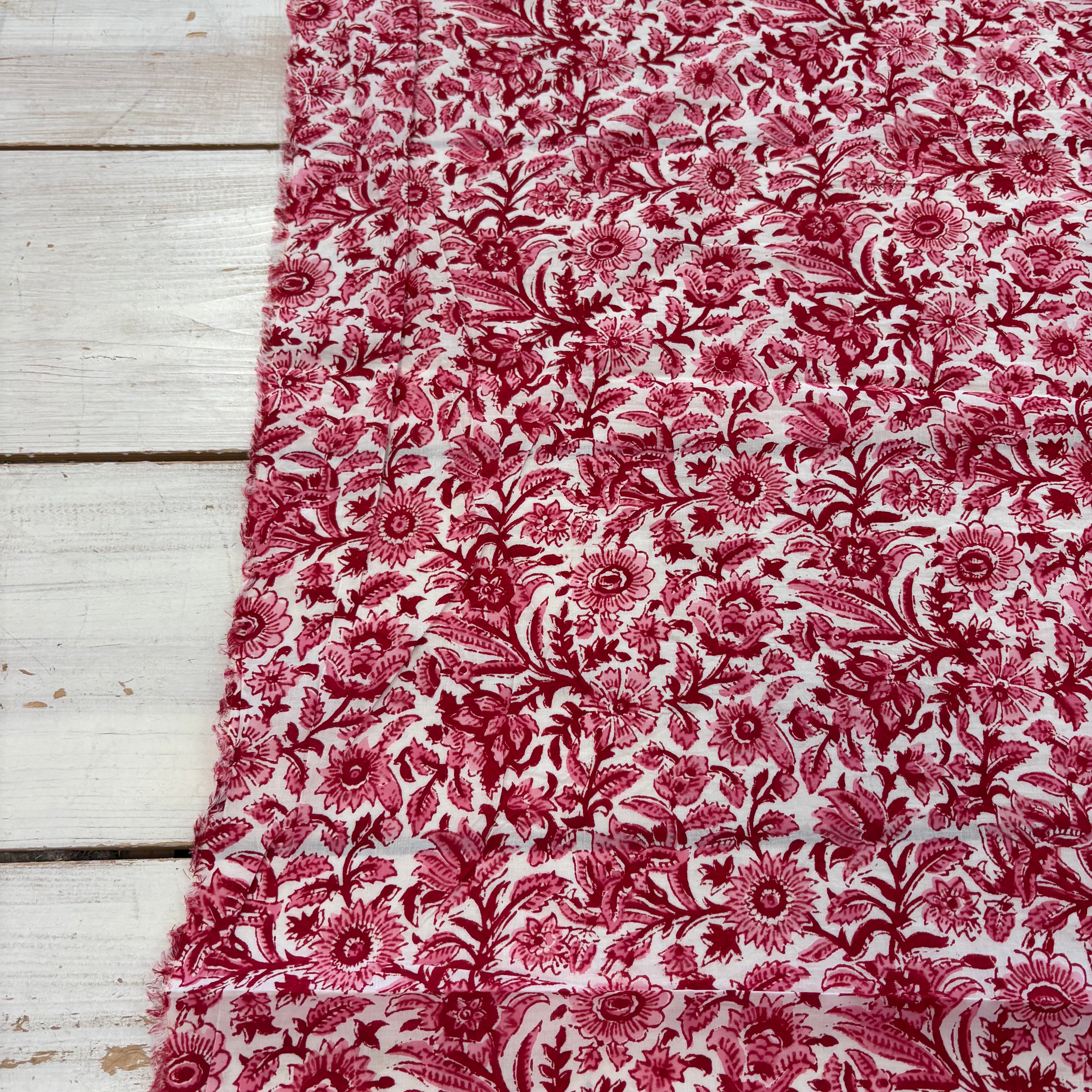 A close-up of a fabric with an all-over floral toile pattern in deep red on a white background, laid flat on a white-washed wooden plank surface.