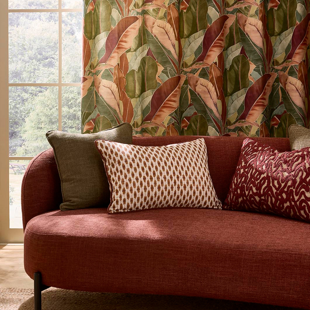 Zafira Burgundy Room Set A styled interior scene showing a terracotta-colored curved sofa with patterned throw pillows in front of a window, with a large tropical leaf patterned curtain hanging on the right side of the window.