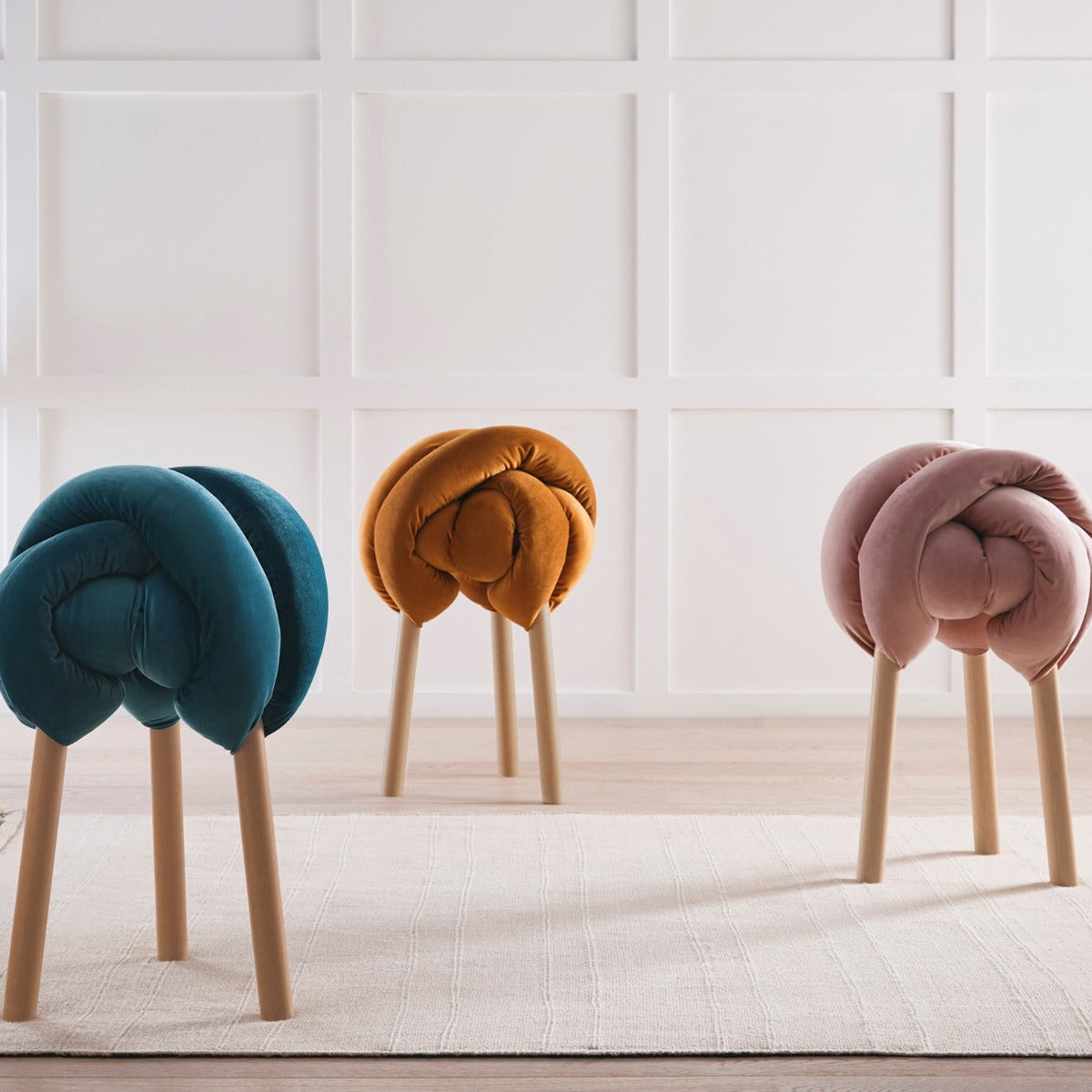 Three unique stools in teal, orange, and pink with wooden legs on a light wooden floor.
